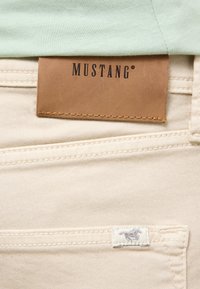 Beige Denim-Jeans mit Mustang-Lederlogo-Patch, ausgestattet mit einem kleinen gestickten Pferd auf der hinteren Tasche und subtiler Textur.