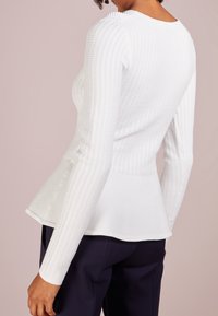 Top bianco a maniche lunghe con orlo peplo, caratterizzato da tessuto testurizzato e scollatura rotonda. Indossato con pantaloni blu scuro.
