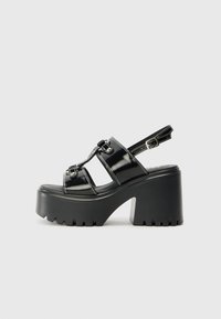 Black platform sandals with glossy patent material, featuring three cross straps and adjustable ankle buckle. Thick, textured sole.