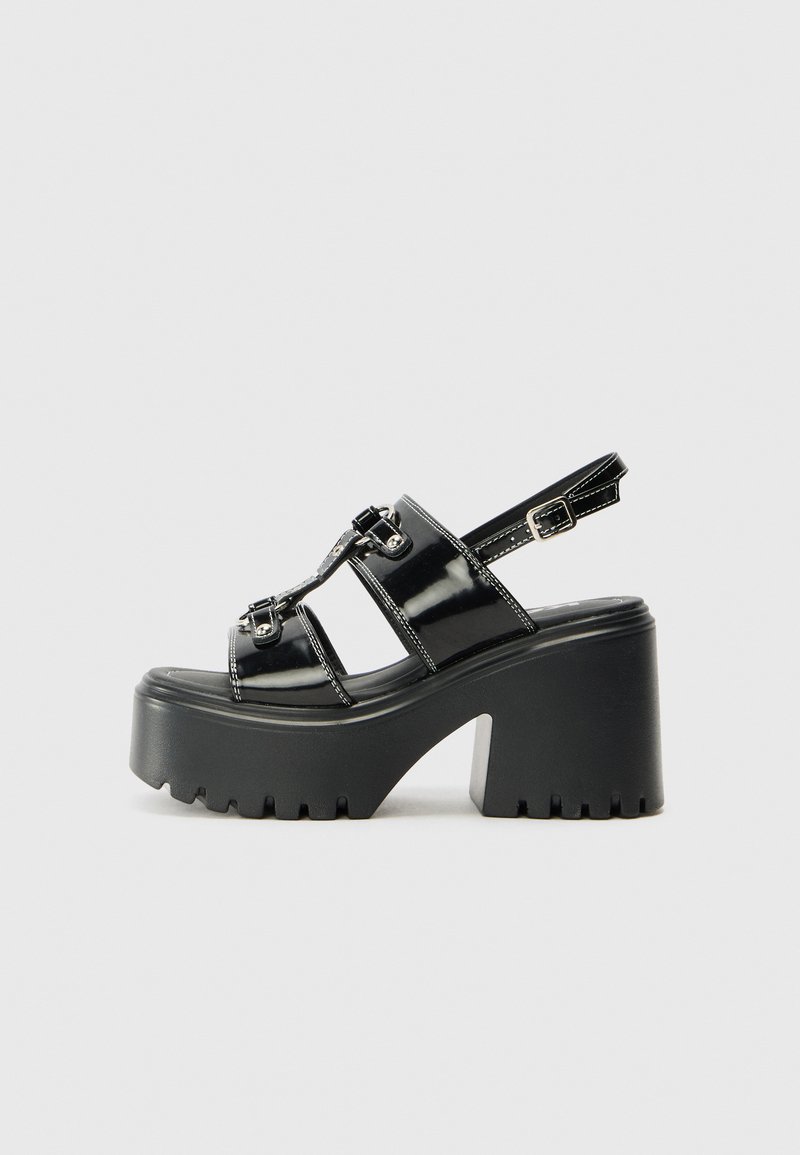 Black platform sandals with glossy patent material, featuring three cross straps and adjustable ankle buckle. Thick, textured sole.