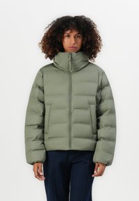 Vrouw in een groene pufferjack met hoge kraag en donkere broek, staande tegen een effen witte achtergrond.