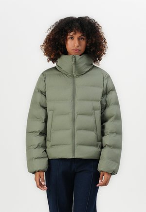 Vrouw in een groene pufferjack met hoge kraag en donkere broek, staande tegen een effen witte achtergrond.