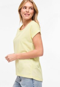 Κοντομάνικη κίτρινη μπλούζα με στρογγυλή λαιμόκοψη, μαλακό βαμβακερό υλικό, άνετη εφαρμογή και ευθεία τελείωμα, σε συνδυασμό με φωτεινό μπλε τζιν παντελόνι.