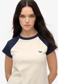 T-shirt à manches courtes avec un corps beige et des manches raglan bleu marine. Présente un petit logo "Super Dry" brodé sur la poitrine.