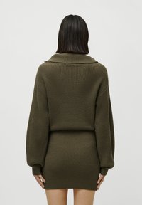 Frau mit glatten, dunklen Haaren, die einen olivgrünen gestrickten Pullover und einen passenden, figurbetonten Rock trägt, von hinten gezeigt.