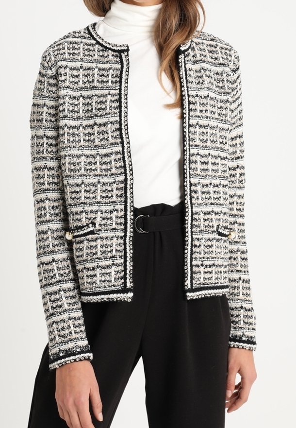 Žena nosící černobílou texturovanou vzorovanou otevřenou bundu přes bílý rolák, doplněnou černými kalhotami s vysokým pasem.