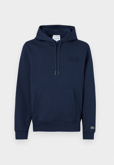 Sweat à capuche bleu marine avec poche avant, capuche à cordon de serrage, petite écusson logo sur la poitrine et logo crocodile sur le poignet de la manche gauche.