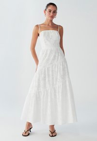 Robe maxi blanche avec un design en couches et des détails brodés. Comporte des bretelles fines et une taille cintrée, associée à des tongs noires.