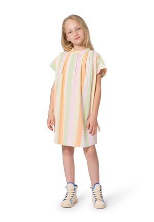 Jeune fille debout, portant une robe ample à manches courtes avec des rayures verticales pastel et des baskets montantes beige avec des accents bleus.