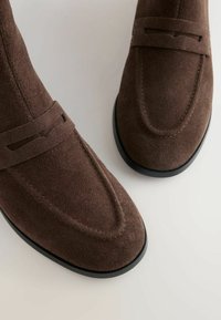 Brązowe zamszowe mokasyny z okrągłym noskiem, z haftowanymi detalami i paskiem na przodzie. Czarna gumowa podeszwa stanowi kontrast.