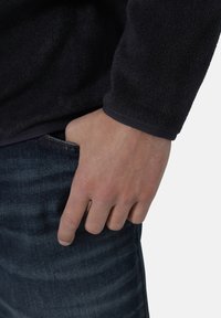 Schwarzer Fleece-Pullover mit strukturierter Oberfläche und engen Bündchen, kombiniert mit dunkelblauen Jeans. Die Hand ruht in der Tasche.