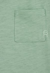 Zelená bavlněná tkanina s texturovaným povrchem obsahuje malou náprsní kapsu a decentně vyšívané písmeno "R" na boku.