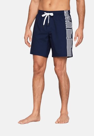 Mann trägt marineblaue Badehose mit weißem Kordelzug und vertikalem weißem Grafikdesign auf der rechten Seite, steht barfuß.