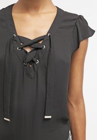 Schwarze Bluse mit V-Ausschnitt und überkreuztem Schnürdesign, silbernen Ösen und kleinen Rüschenärmeln. Glatte Textur und weicher Stoff.