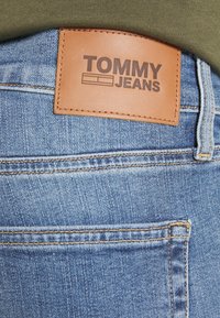 Siniset farkut, joissa on nahkainen tagi, jossa lukee "TOMMY JEANS". Ominaisuuksiin kuuluvat ompeluyksityiskohdat ja klassinen viisi-taskuinen muotoilu.