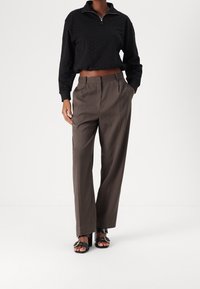 Haut texturé noir à fermeture éclair associé à un pantalon marron taille haute. Le mannequin porte des sandales à talons noires avec des accents dorés.