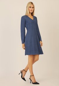 Vestito lavorato a maglia blu con scollatura a V, presentante maniche lunghe e design a trecce testurizzate, che si allarga dolcemente all'orlo. Scarpe nere con tacco.