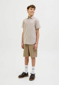 Chemise beige à manches courtes, shorts beiges, et chaussures noires avec des semelles texturées. Des chaussettes blanches avec un texte imprimé sont visibles.