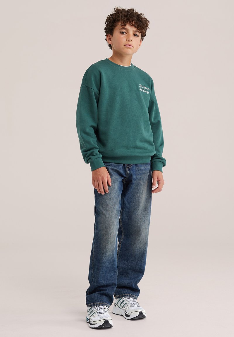 Garçon aux cheveux bouclés portant un sweat-shirt vert, un jean bleu et des baskets blanches, debout devant un fond uni.