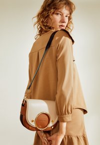 Una donna con i capelli ricci indossa un vestito beige, mentre porta una borsa a tracolla in pelle bicolore beige e marrone con dettaglio in oro a cerchio e catena.