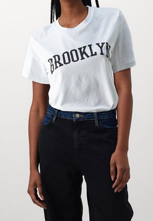 White cotton t-shirt featuring "BROOKLYN" in bold black letters, paired with dark denim pants, showcasing a casual fit and smooth texture.