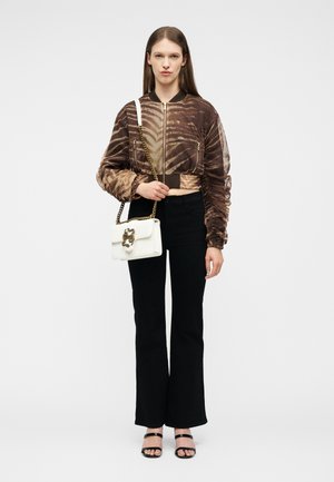 Jonge vrouw die een bruin patroonritsjack draagt, zwarte wijde broek, zwarte sandalen, en een witte handtas met gouden kettingriem vasthoudt.