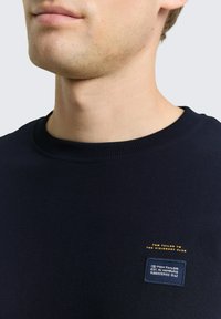 Granatowa bluza z prążkowanym dekoltem, ozdobiona złoto-niebieskimi haftowanymi logo na piersi oraz miękkim, teksturowanym materiałem.