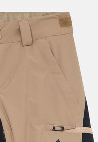 Beige utomhusbyxor med snedställda framfickor, svarta sidopaneler, en ficka med dragkedja och rem, bälteslök, samt justering med kardborreband i midjan.