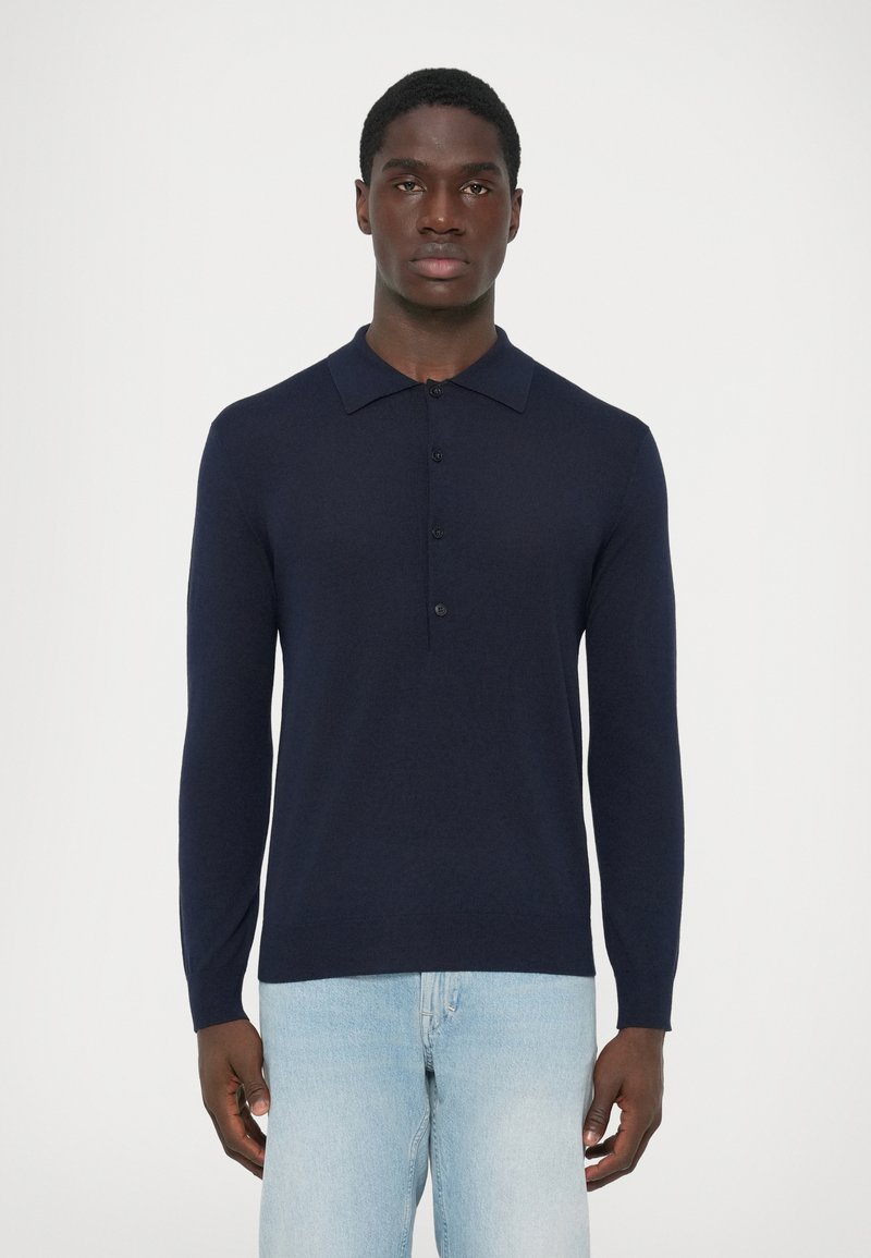 Uomo con una maglietta polo a maniche lunghe blu navy e jeans azzurri, in piedi contro uno sfondo bianco neutro.