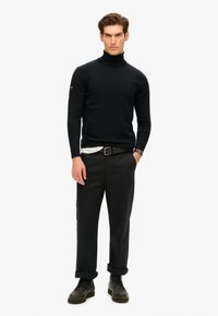 Maglione a collo alto nero con una texture liscia, abbinato a pantaloni scuri e scarpe nere con lacci. Design minimalista; vestibilità casual.
