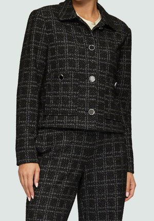 Femme portant un blazer à carreaux noir et blanc avec des boutons métalliques et un pantalon taille haute assorti, debout devant un fond uni.