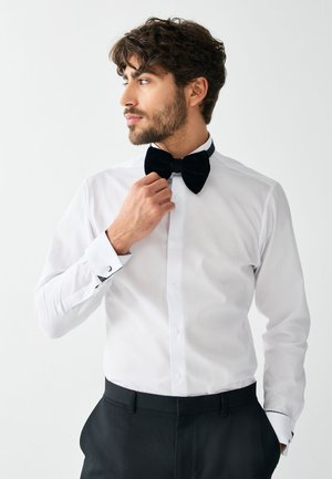 Mand iført hvid skjorte og sort butterfly justerer sin butterfly og ser til siden mod en enkel, lys baggrund.