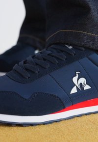 Scarpe atletiche navy con materiali scamosciati e sintetici, dettagli bianchi e rossi, punta arrotondata, suola testurizzata e logo prominente sul lato.