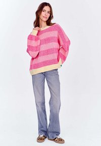 Różowy i kremowy sweter w paski z luźnym krojem, bufiastymi rękawami i prążkowanym dekoltem. Noszony z jasnoszarymi dzwonami i wzorzystymi butami.