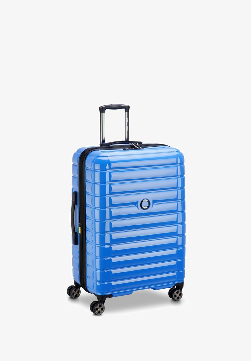 Valise rigide bleue avec texture rainurée, fermeture éclair noire et poigné de traction. Dispose de quatre roues pour la mobilité et d'un accent de logo circulaire.