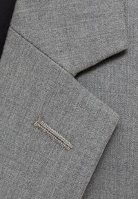 Tissu de blazer gris montrant un tissage texturé, avec un détail de couture décoratif sur le revers et une ouverture de poche à l'avant.