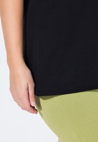 Maglietta nera in cotone con orlo e spacchi laterali, abbinata a leggings aderenti di un verde chiaro, che mostrano una texture liscia e bordi puliti.