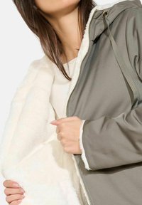 Olivgrüne Zip-Jacke mit weißem Fleece-Futter, ausgestattet mit Kapuze und Metallreißverschluss. Weiche Textur; sichtbare Nähte und Details mit Kordelzug.