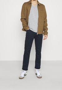 Braune Zip-Jacke, graues V-Ausschnitt-T-Shirt, marineblaue Hose und weiße Sneakers mit schwarzen Akzenten. Die Jacke hat eine Brusttasche und Bündchen.