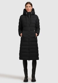 Palton lung negru tip puffer cu glugă, având secțiuni matlasate, fermoar frontal și două buzunare laterale. Purtați peste colanți negri și cizme.