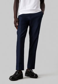 Pantaloni blu navy in tessuto leggero con vestibilità dritta e texture liscia, abbinati a scarpe nere lucide. Linee pulite e design minimal.