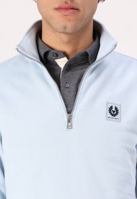 Uomo che indossa un maglione azzurro chiaro con zip a 1/4 sopra una polo grigia scura con bottoni, il maglione presenta una patch del logo Belstaff sul petto.