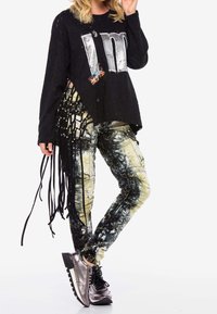 Camiseta oversized negra de manga larga con un gráfico metálico que dice "Soy", combinada con pantalones ajustados multicolores que presentan patrones abstractos y detalles de flecos.