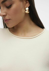 Boucle d'oreille en métal doré, brillante, avec un design lisse et arrondi, portée par un mannequin sur un haut en tissu couleur crème.
