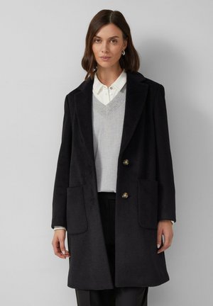 Manteau en laine noire au design structuré, avec un col à revers, deux poches avant et deux boutons. Porté par-dessus un pull gris et une chemise.