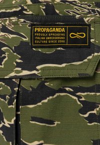 Tessuto mimetico con sfumature di verde e nero, caratterizzato da un'etichetta nera con testo giallo che recita "PROPAGANDA SPREADING PROUDLY UNDERGROUND ITALIAN CULTURE DAL 2006."