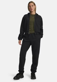 Black zippered hoodie with kangaroo pockets and cropped design over a dark green fitted shirt. Black sweatpants with elastic cuffs. Black sneakers.