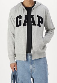 Grauer Hoodie mit Reißverschluss und schwarzem "G.A.P."-Logo, mit Kordelzugkapuze, Fronttaschen sowie gerippten Bündchen und Saum. Wird über einem schwarzen T-Shirt getragen.