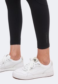 Πόδια που φορούν μαύρα κολάν Calvin Klein και λευκά αθλητικά παπούτσια Adidas με κορδόνια σε απλό φόντο.