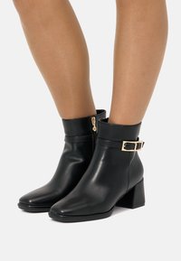 Botines negros de piel suave, con puntera afilada, tacón medio y un detalle de hebilla dorada en el tobillo.